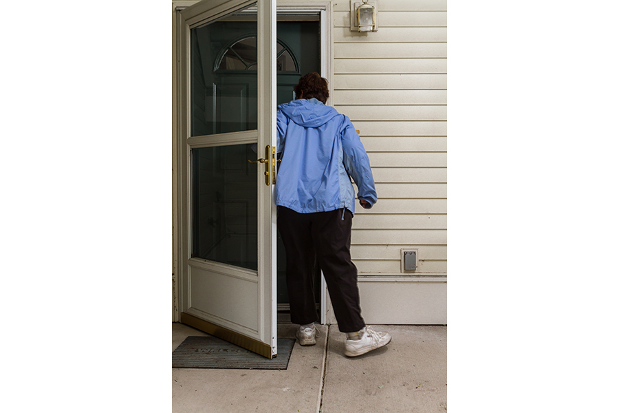 A woman with orthopedic shoes and a blue jacket steps through an open screen door to enter a green front door of a home with a white facade.